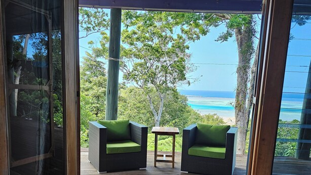 An open wooden deck with two green cushioned chairs and a small table, leading to a scenic view of the ocean and trees.
