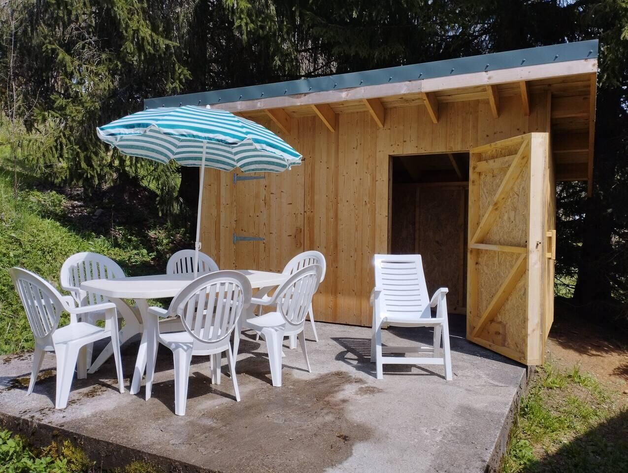 An outdoor wooden structure with a concrete patio, white plastic chairs, a table, and a striped umbrella.