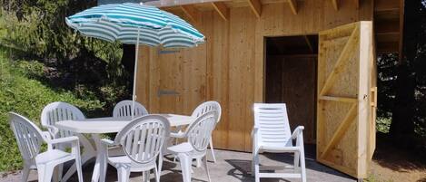 An outdoor wooden structure with a concrete patio, white plastic chairs, a table, and a striped umbrella.