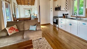 A spacious living area with a wooden ceiling, a sofa with decorative pillows, a dining table, and a kitchen with white cabinets and a microwave.
