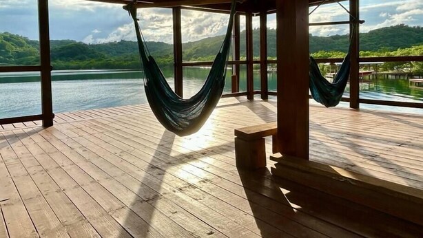 A wooden deck with two hammocks hanging from the ceiling, overlooking a calm lake and lush green hills.