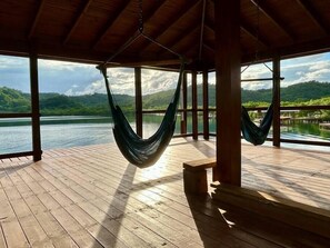 Een houten terras met twee hangmatten aan het plafond, met uitzicht op een kalm meer en glooiende groene heuvels.