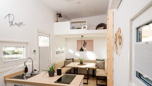 Een compacte keuken met een spoelbak, aanrecht en een klein eiland. Er is een zithoek met een bank en een tafel, en daarboven een slaapverdieping met een bed en opbergruimte.