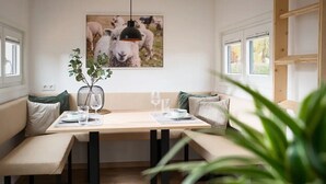 Een eetgedeelte met een houten tafel gedekt voor twee personen, bankjes met kussens, een hangende hanglamp en een lijst met een afbeelding van schapen aan de muur.