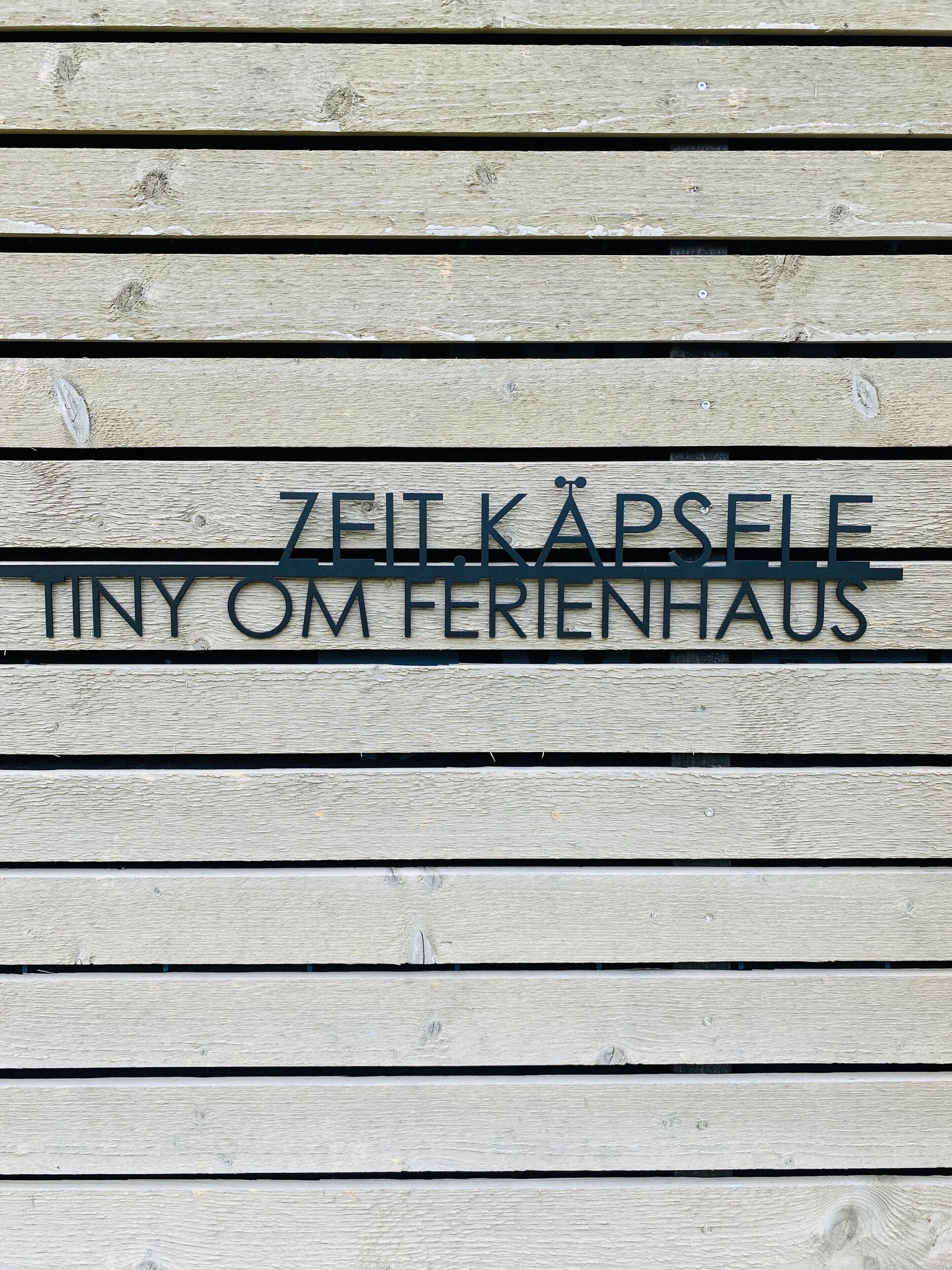 A wooden sign with the text 'ZEIT KAPSELE TINY OM FERIENHAUS'.