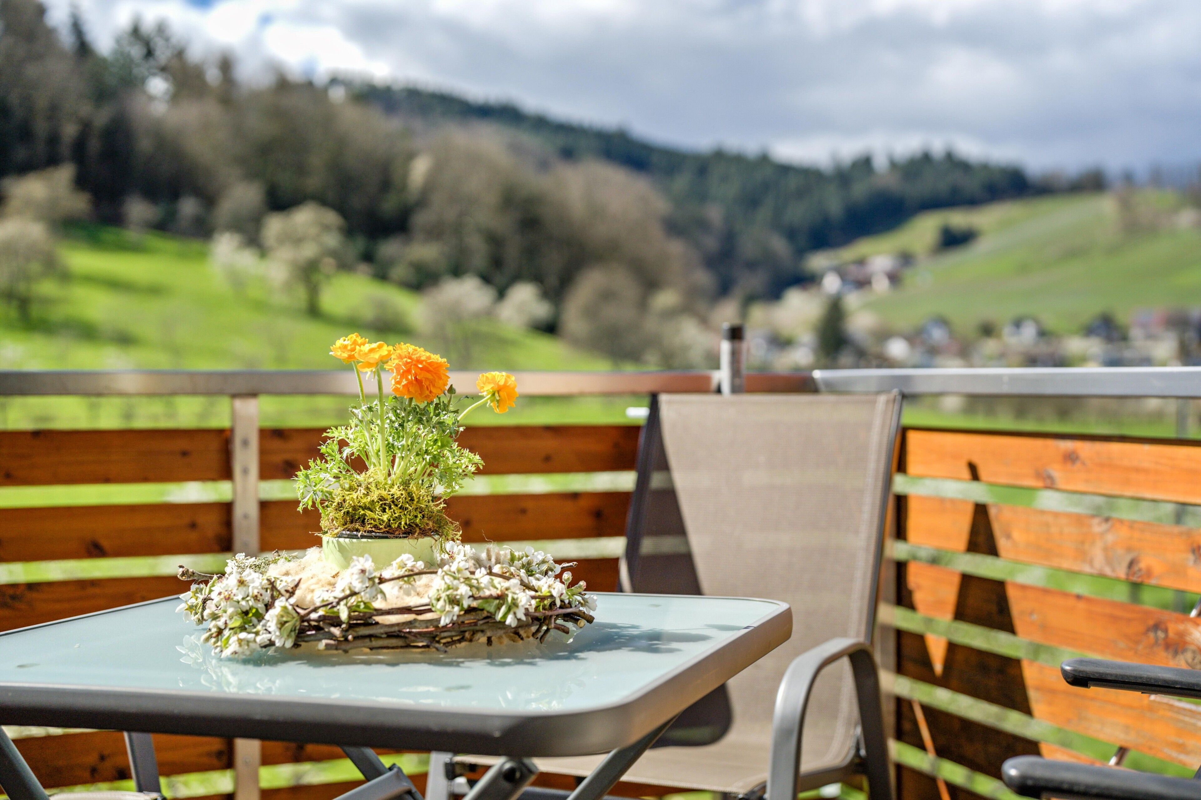Mesa con planta en maceta y flores, una silla y una valla de madera con vistas a colinas verdes y casas.