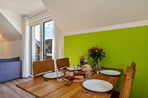 Un coin repas avec une table en bois dressée pour deux personnes, un canapé et une vue sur un bâtiment à l’extérieur.