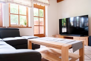 Olohuoneessa on sohva, puinen pöytä, televisio ja ikkuna, josta avautuu näkymä vehreään maisemaan.