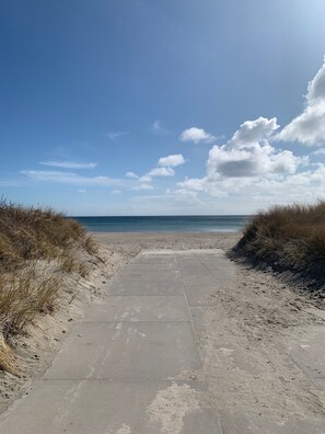Ein gepflasterter Weg führt zu einem Strand mit Dünen links und rechts.