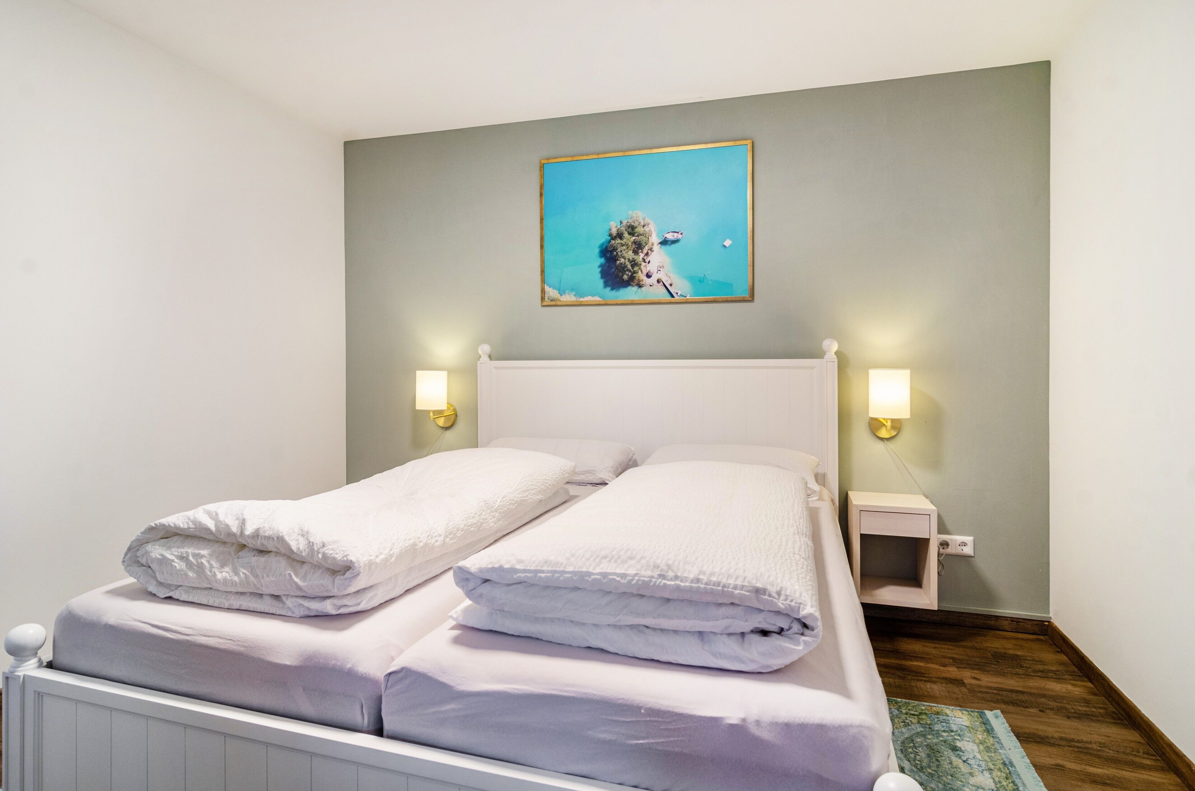 En pænt redt seng med hvide sengetæpper, et lille natbord med en lampe og et billed af et kystlandskab hængt på væggen.
