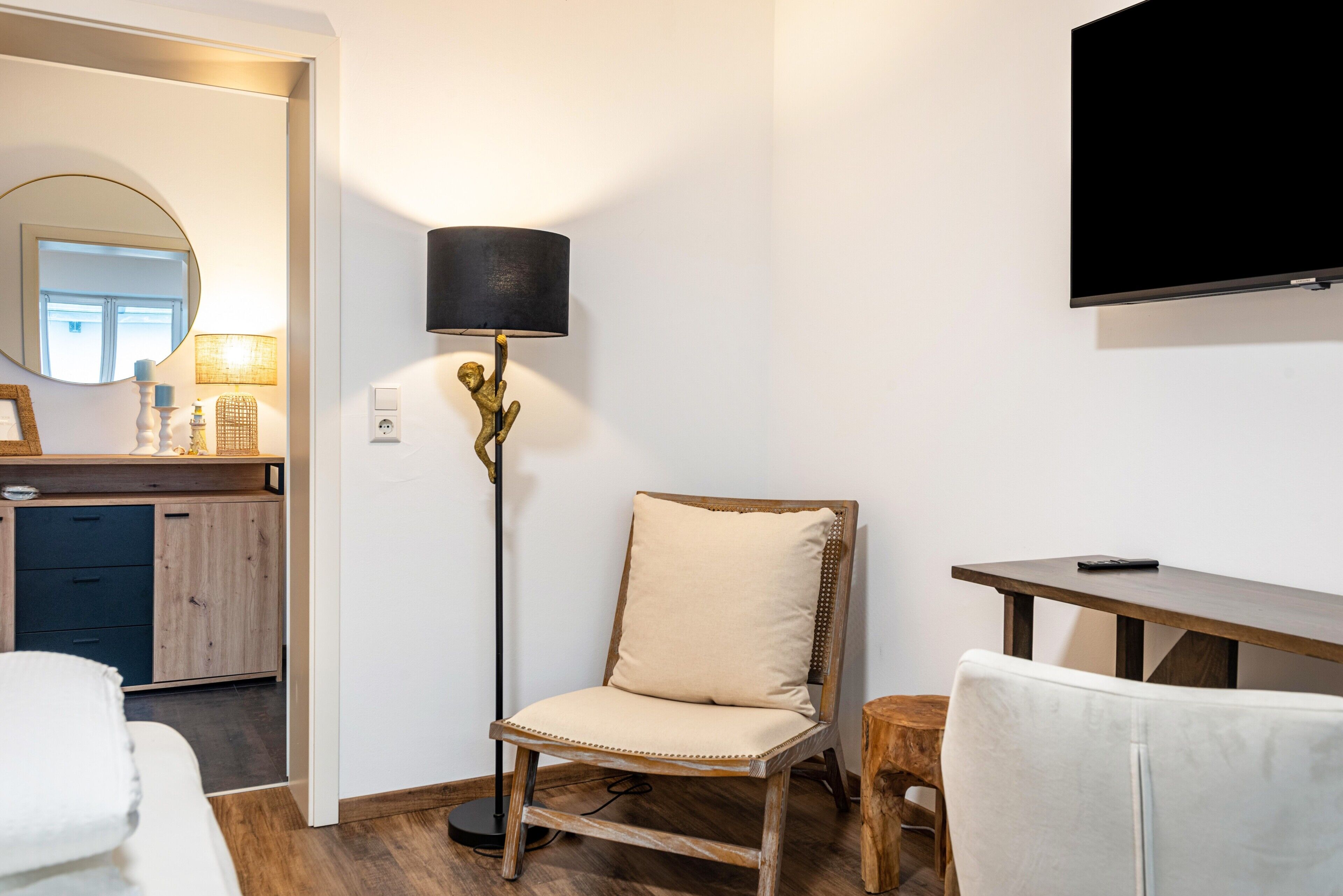 A room with a wooden floor, a chair with a cushion, a small wooden table, a floor lamp, a mirror, and a television mounted on the wall.