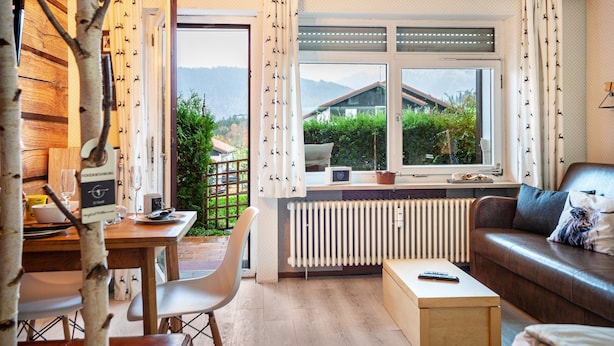 A cozy living room with a wooden dining table, a sofa, and a view of greenery and mountains through the window.