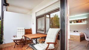 A patio with a wooden table and two chairs, a window, and a bed in the adjacent room.