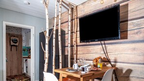 A dining area with a wooden table, white chairs, a television mounted on the wall, and a kitchen area in the background.