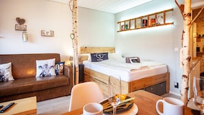 A cozy room with a wooden bed, a sofa with cushions, a small table with a glass and a plate, and a wall shelf with books.