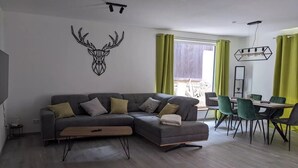 Een moderne woonkamer met een hoekbank, een eethoek met tafel en stoelen, en een groot raam met gordijnen.