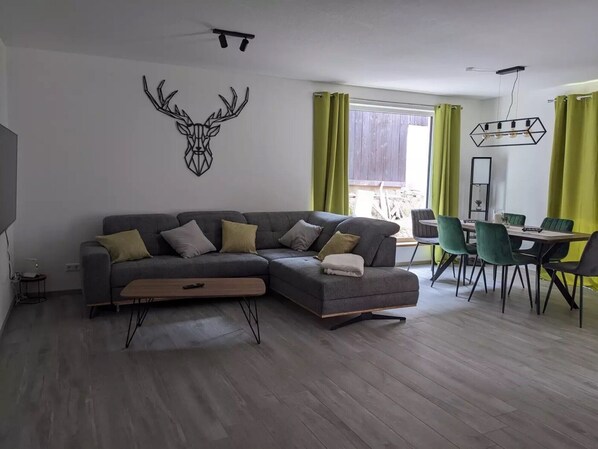 A modern living room with a sectional sofa, a dining area with a table and chairs, and a large window with curtains.