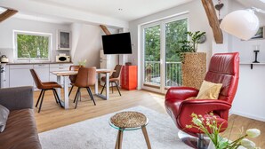 A modern interior with a dining area, a red leather chair, a flat-screen TV, and a balcony with a view of greenery.