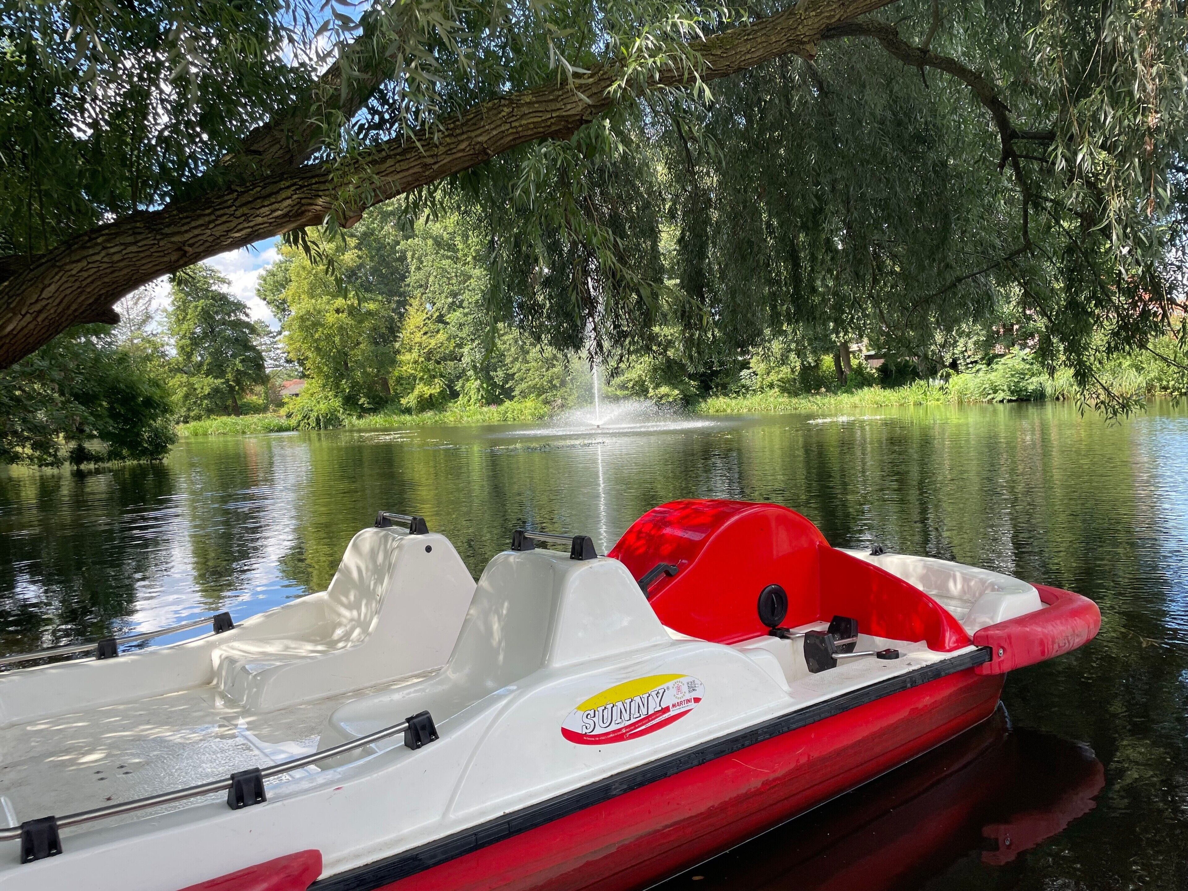 Um barco com esquema de cores vermelho e branco está atracado perto de um rio calmo, cercado por vegetação exuberante e árvores.