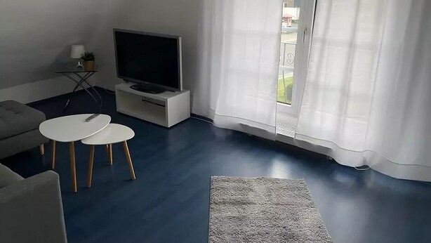 Un salon avec un téléviseur à écran plat posé sur un meuble blanc, un canapé gris, deux petites tables rondes blanches et un tapis gris.