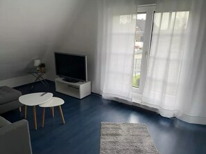 A living room with a flat-screen TV on a white stand, a grey sofa, two white round tables, and a grey rug.