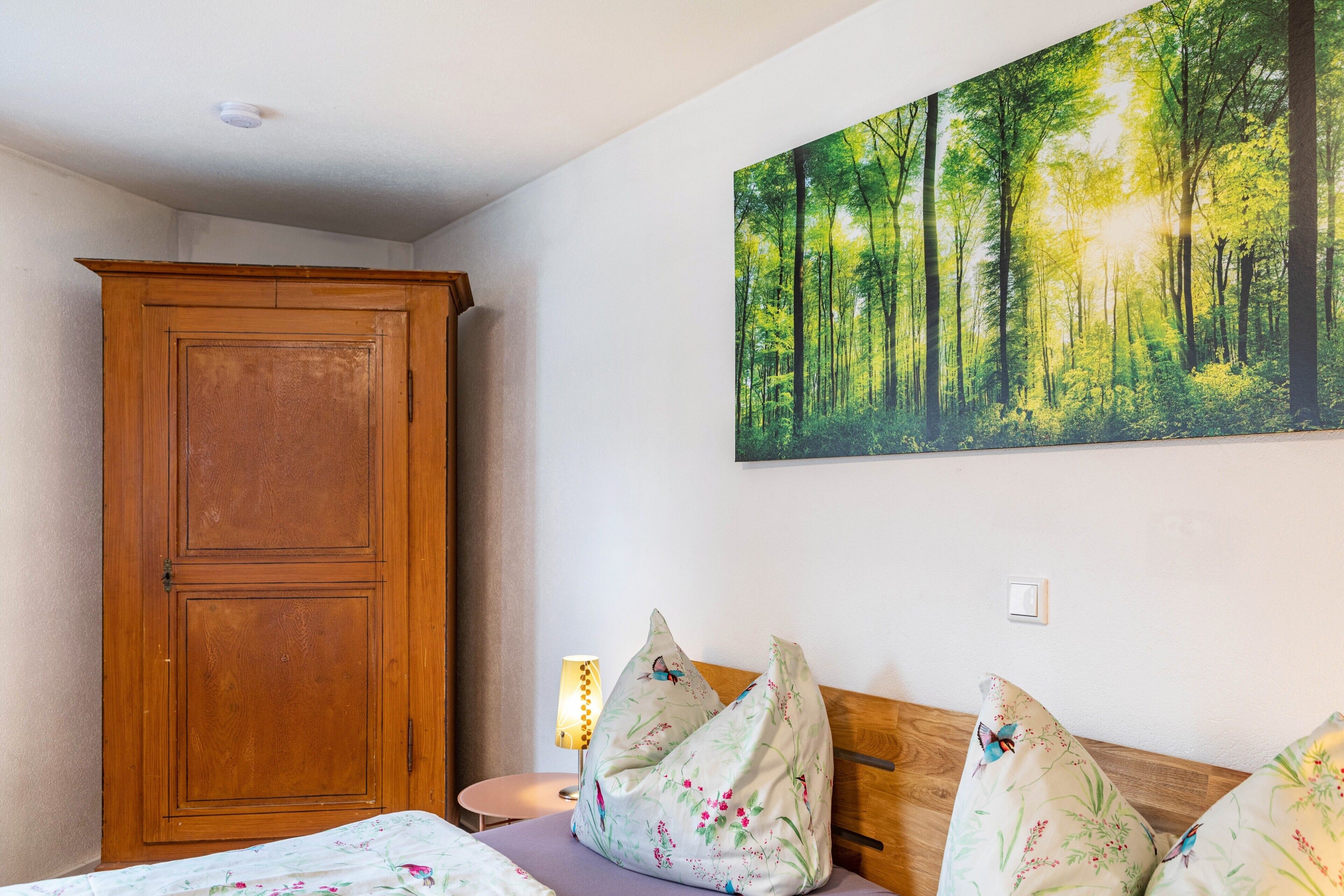 Une chambre à coucher avec un lit, une armoire en bois, un tableau représentant une forêt accroché au mur et une lampe de chevet.