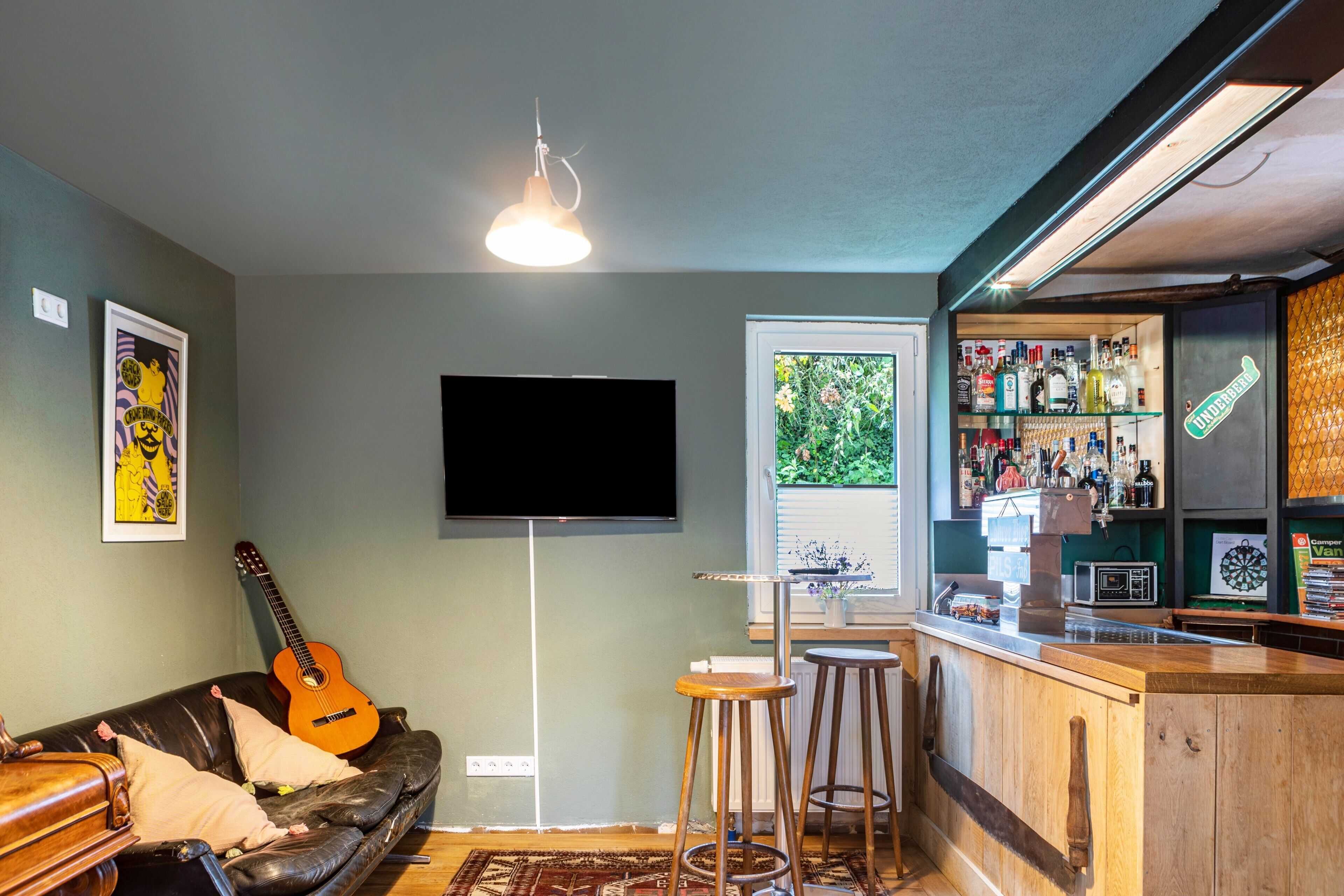 Un salon avec un téléviseur, une guitare, un coin bar avec des bouteilles et une fenêtre donnant sur un espace verdoyant.