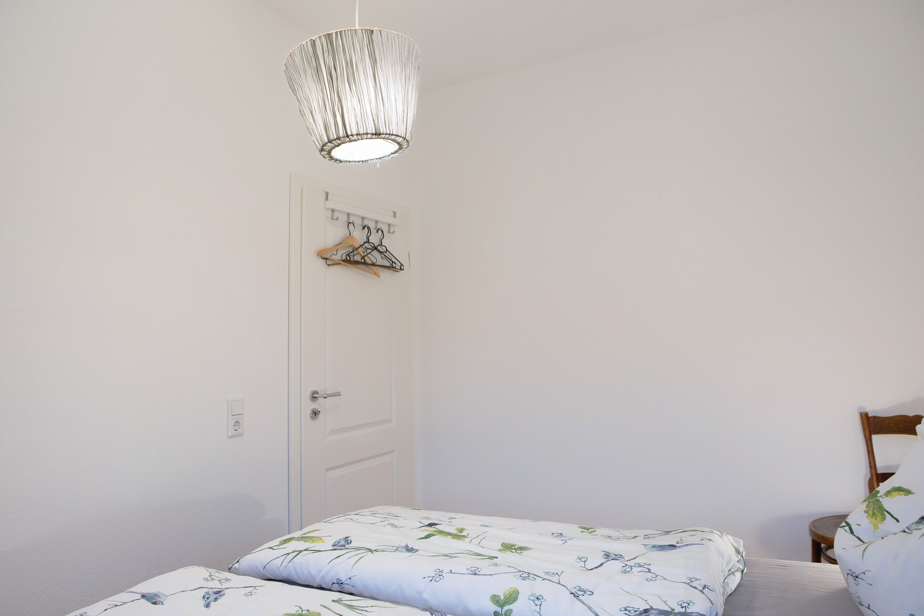 Ein Schlafzimmer mit zwei Betten, einer Hängelampe, einer Tür mit Kleiderhaken und einem Sessel.