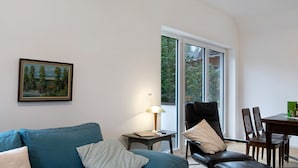 Un salon avec un canapé, un fauteuil, une table et une lampe. Il y a une fenêtre qui donne sur des arbres.