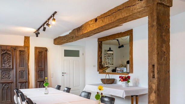 Un espace repas avec une longue table, des chaises, des poutres en bois, une porte blanche et un miroir au mur.