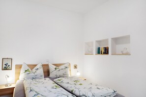 A bedroom with a bed, bedside tables, a nightstand, a wall-mounted shelf with books, and a framed picture.