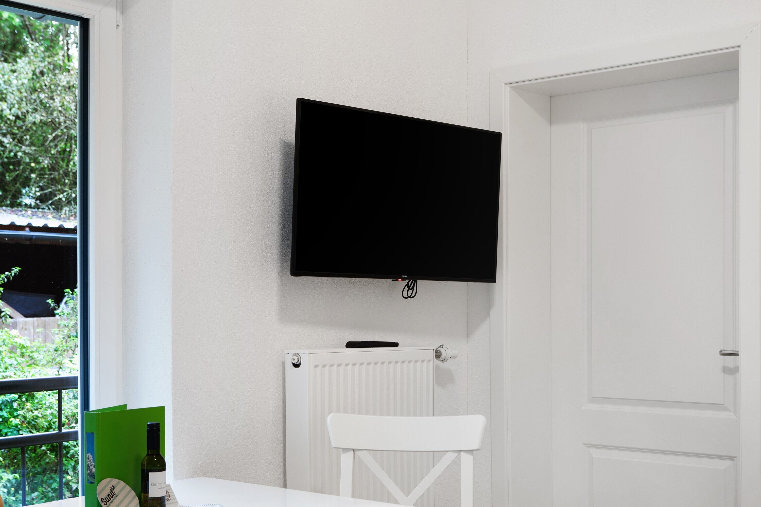 A room with a white radiator, a white chair, a television mounted on the wall, and a window with a view of greenery.
