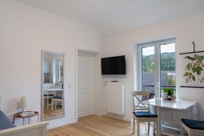 Une chambre moderne avec un plancher en bois, un bureau blanc, un téléviseur fixé au mur et une grande fenêtre donnant sur des arbres et des immeubles.