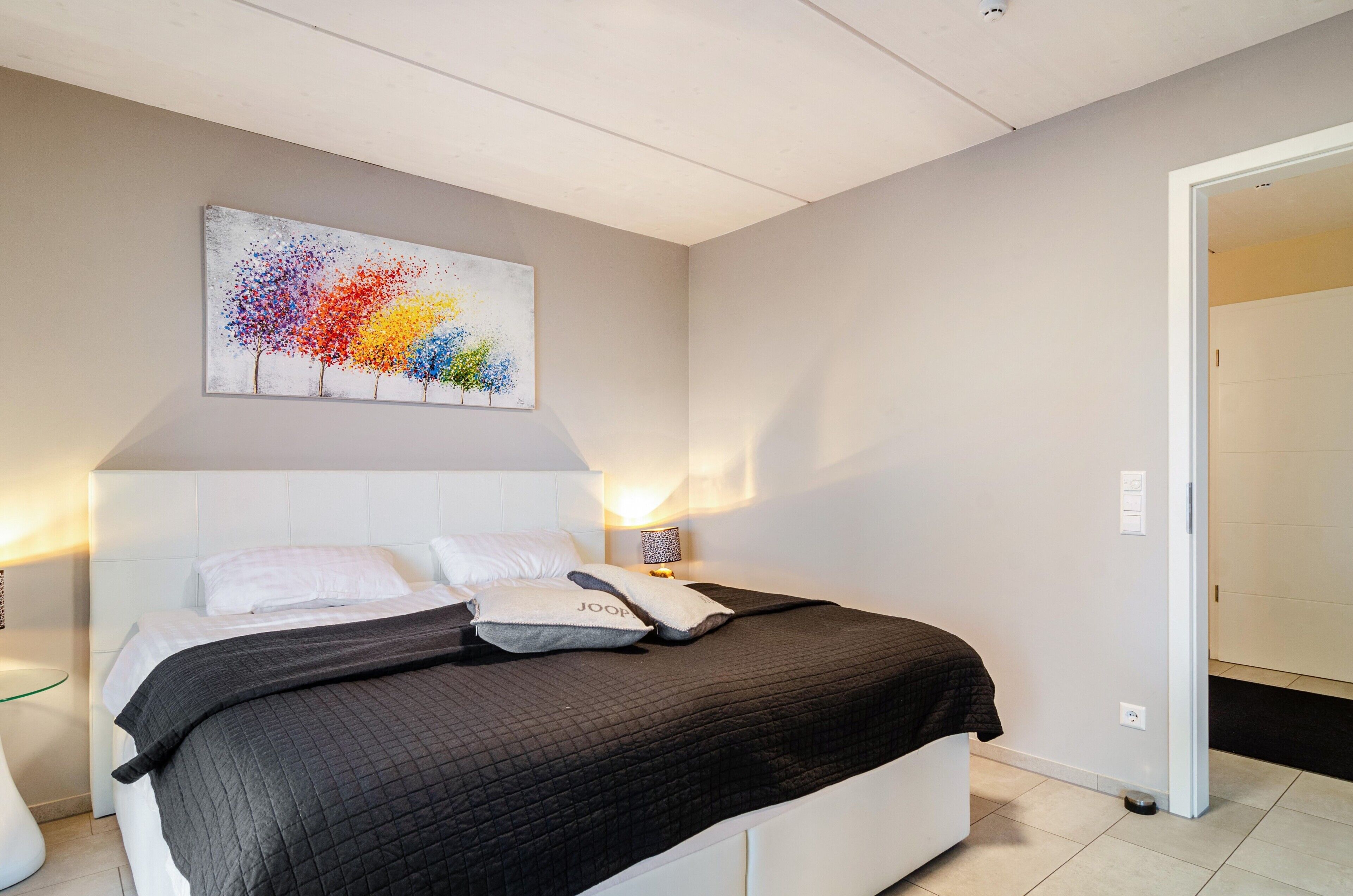 A bedroom with a bed, a wall-mounted artwork, bedside lamps, and a door leading to another room.