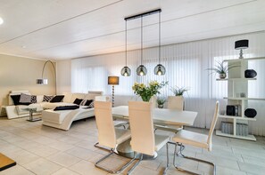 Un salon moderne avec un canapé blanc, une table à manger avec des chaises, une lampe sur pied et une bibliothèque.