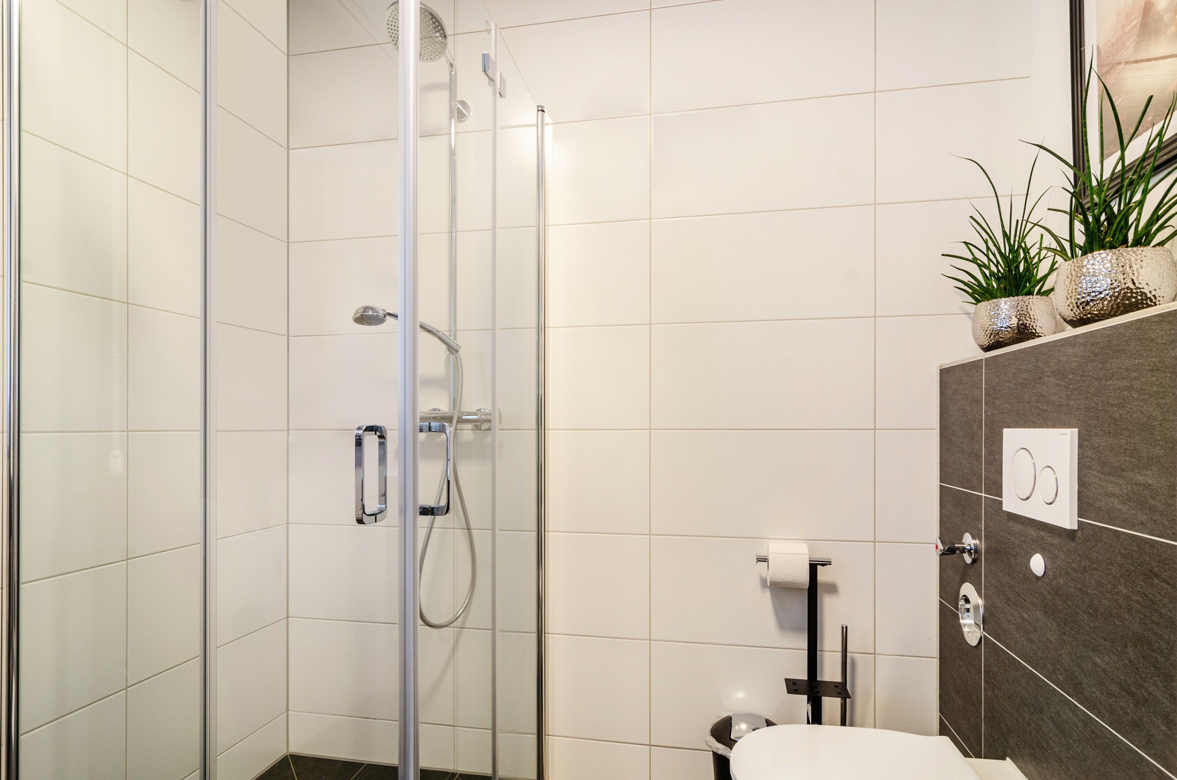 Un baño moderno con cabina de ducha de vidrio, inodoro y tocador con plantas decorativas.