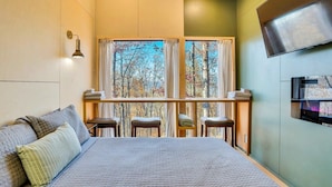 A bedroom with a large bed, a window with a view of trees, a mounted TV, and a small seating area with stools.