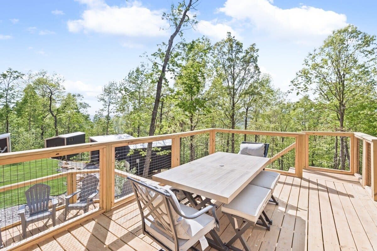 Terraza de madera con una mesa y sillas, rodeada por barandilla de madera y vistas a árboles.