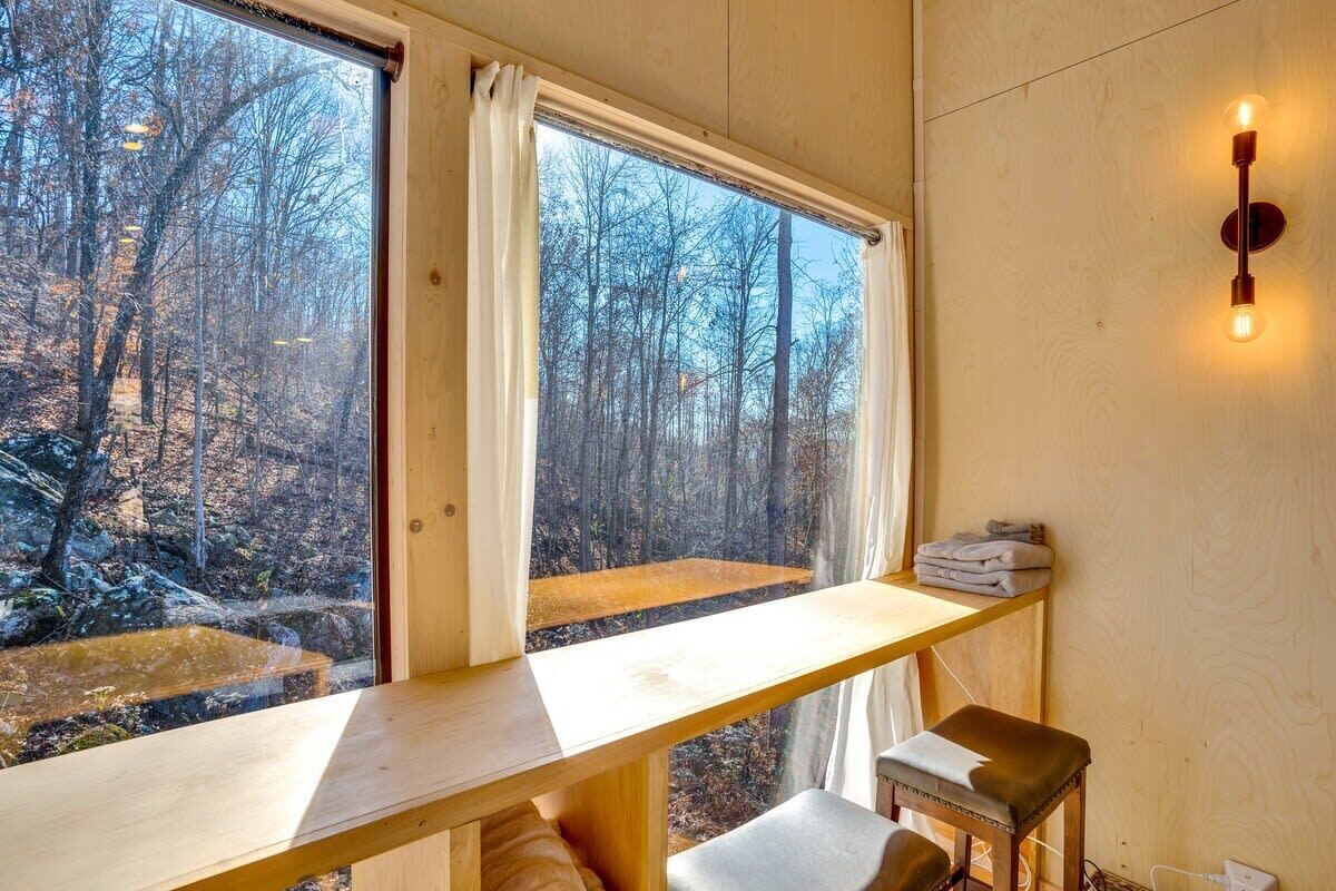 A room with a wooden desk, a bench, and a view of a forest through large windows.