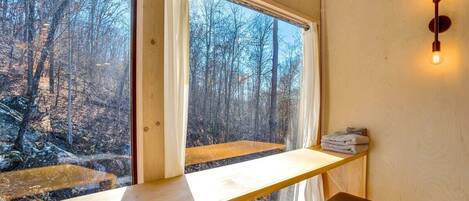 A room with a wooden desk, a bench, and a view of a forest through large windows.