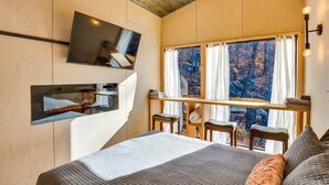 A bedroom with a bed, a TV mounted on the wall, a window with curtains, and a small table with chairs.