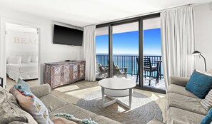 En moderne stue med stort TV, sofa, sofabord og balkon med havudsigt.