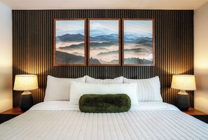 A neatly made bed with white linens, a green throw pillow, and two bedside lamps. Above the bed, there are three framed landscape paintings on a wooden panel wall.