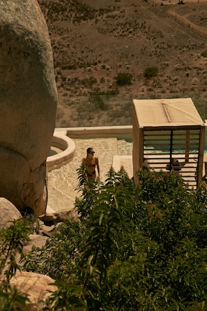 A person in a bikini walking towards a pool area with a large rock and a wooden structure nearby.