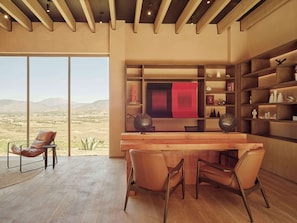 A spacious room with a large window offering a view of the desert landscape, a wooden desk with chairs, and built-in shelving with various decorative items. - Banyan Tree Veya Valle De Guadalupe (Valle de Guadalupe)