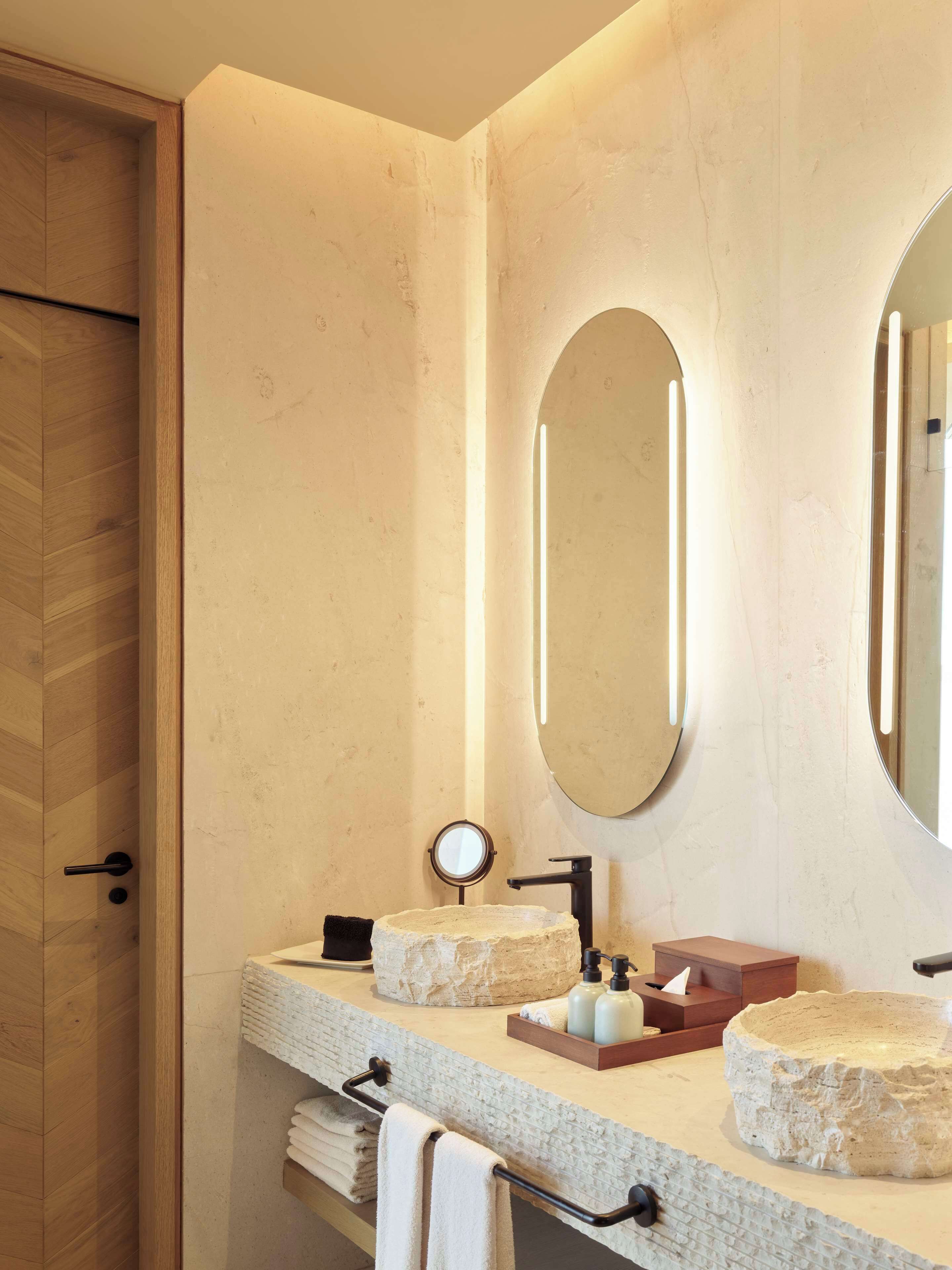 Un baño con puerta de madera, un espejo redondo, un lavabo con cuenca de piedra y un perchero para toallas.