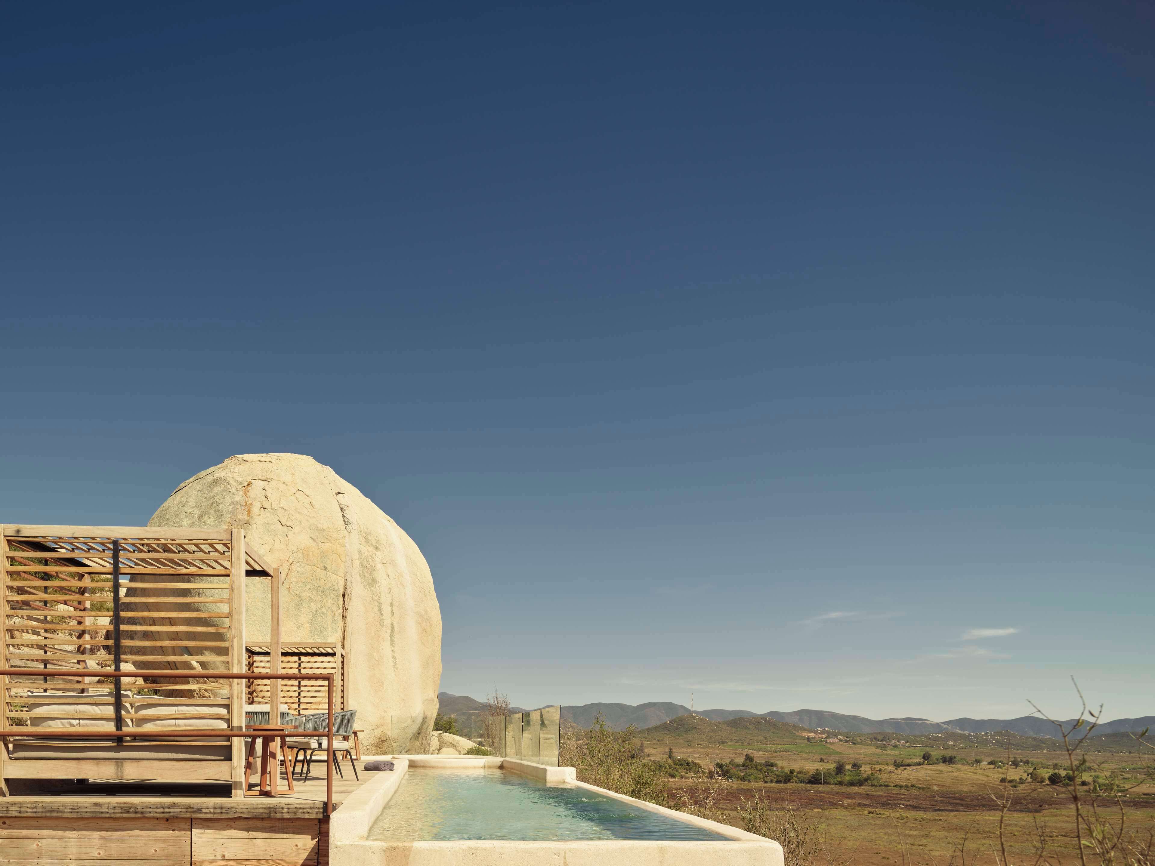 A pool area with a large rock structure, wooden seating, and a view of distant hills.