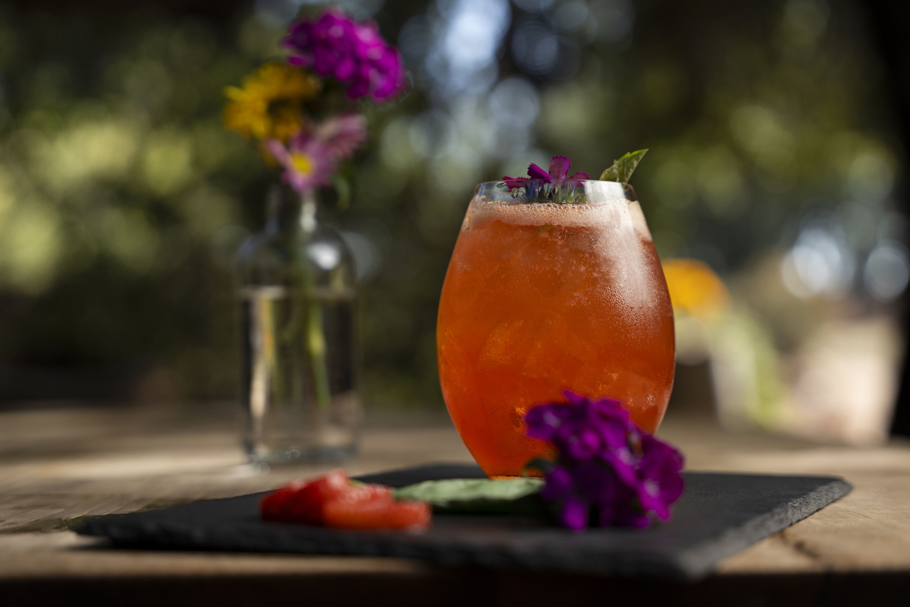 A glass of orange-colored drink with a garnish on a black coaster, accompanied by a small vase of flowers and a strawberry.