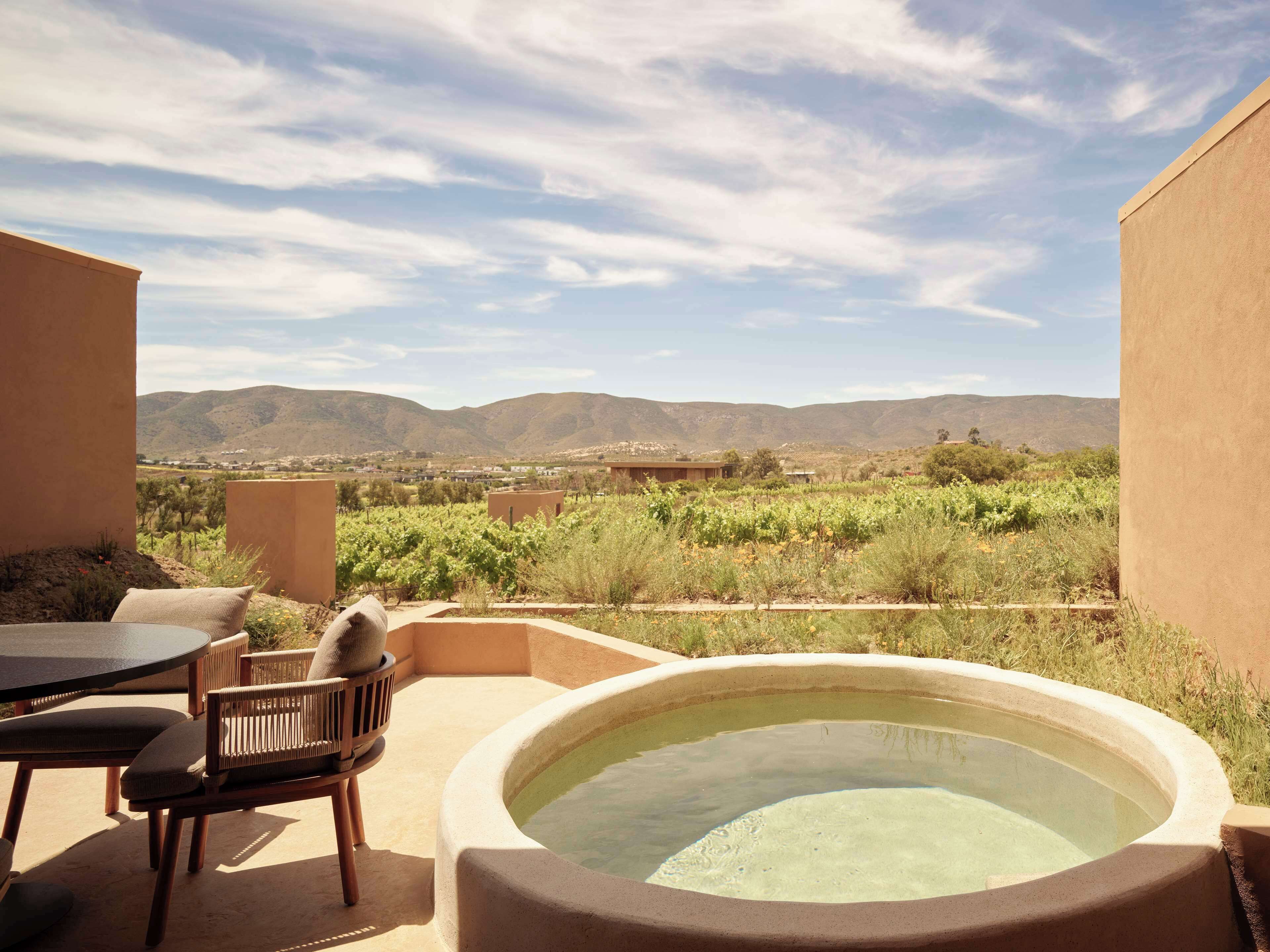 A patio with a hot tub, a table, and chairs, overlooking a vineyard and mountains.