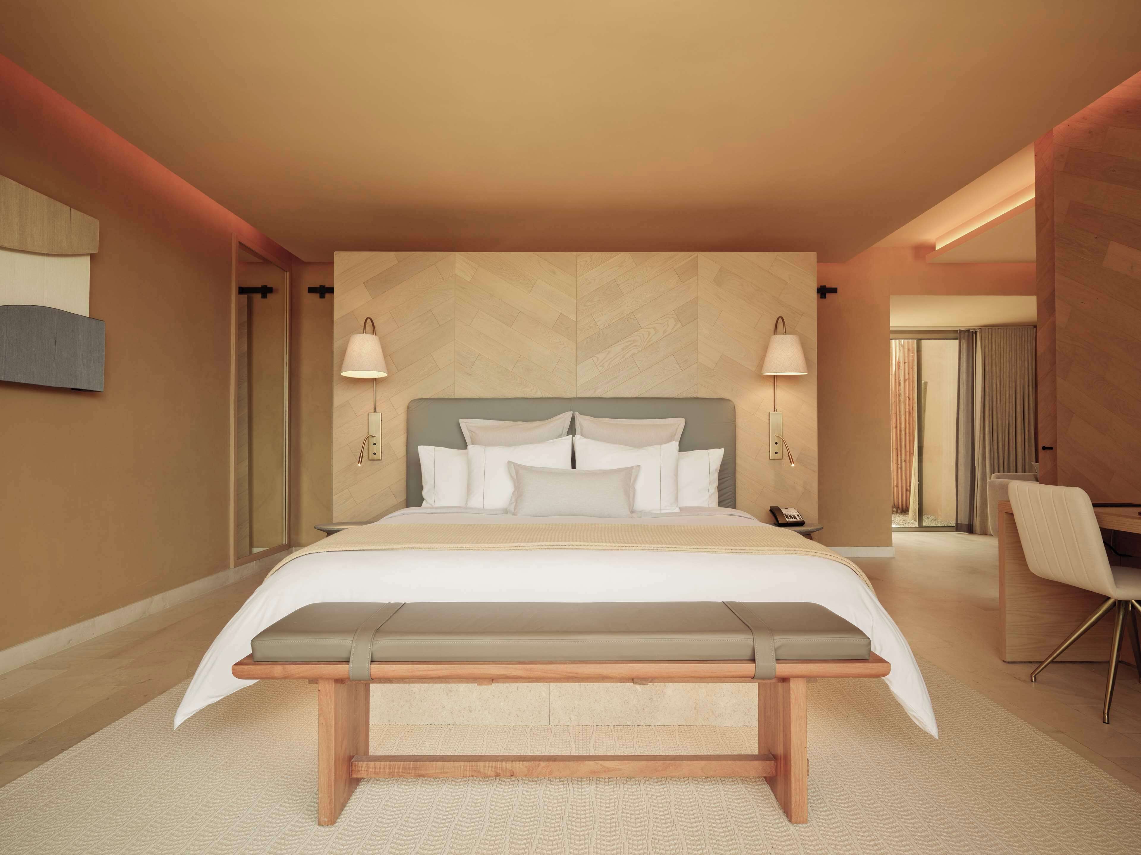 Habitación de hotel moderna con una cama grande, lámparas de noche, un banco y un escritorio con silla.